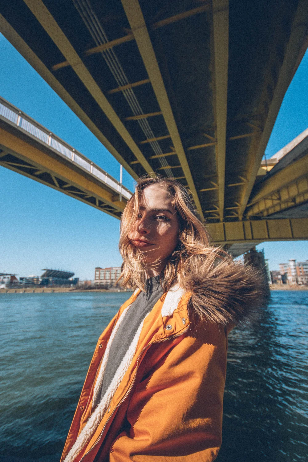 Sunlit portrait of a person in an orange coat beside water beneath a bridge